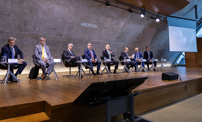 Foto mostra oito magistrados, todos de terno e gravata, sentados em palco de auditório de madeira, tendo à frente de cada mesa baixa com garrafa de água e copo e placa de indicação de seus nomes. Ao fundo, a projeção da palavra abertura, indicando tratar-se da mesa de abertura de seminário