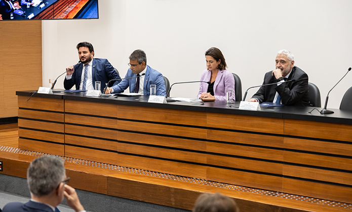 Foto de bancada em auditório com quatro palestrantes - três homens trajando terno e uma mulher de blazer lilás,  sentados à frente de microfones instalados no tampo. Eles estão falando, cada um em seu momento, para uma plateia presente ao auditório