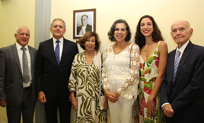 À esquerda dois homens, um de terno cinza e outro de terno azul marinho, estão posicionados ao lado de três mulheres, duas senhoras, uma de vestido estampado de folhagem e outra de vestido branco e a terceira com vestido de estampa floral vermelho e verde sobre fundo branco. Ao lado delas está outro homem de terno azul marinho. Todos posam para uma foto com o retrato de um homem homenageado  na parede 