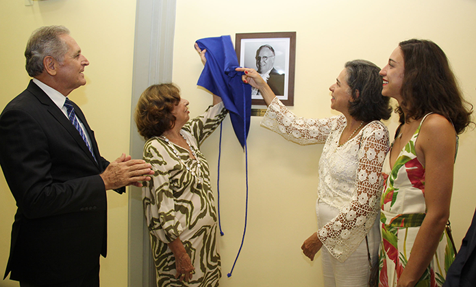 Na foto aparecem três mulheres - uma de vestido de fundo branco com estampa verde; outra de vestido branco e a mais jovem de vestido branco com estampa de floral vermelho e verde - e um homem de terno azul marinho, camisa branca e gravata azul listrada. As mulheres estão descerrando a foto pendurada na parede de um homem homenageado que estava coberta com pano azul. O homem de terno aplaude a ação. Todos estão em pé.  