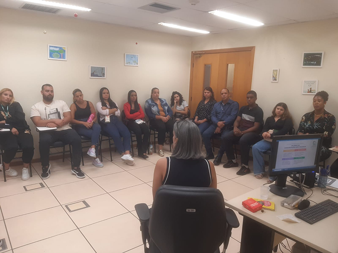 A imagem mostra um ambiente de reunião. Há um grupo de pessoas sentadas em círculo participando de uma palestra. No primeiro plano, uma mulher de cabelos grisalhos está sentada de costas para a câmera, apresentando o tema. À frente dela, há uma mesa com um computador, que exibe uma tela com um slide de apresentação. O ambiente é uma sala fechada, com paredes claras decoradas com alguns quadros e uma porta de madeira ao fundo. Os participantes estão vestidos de forma casual e parecem atentos à atividade.