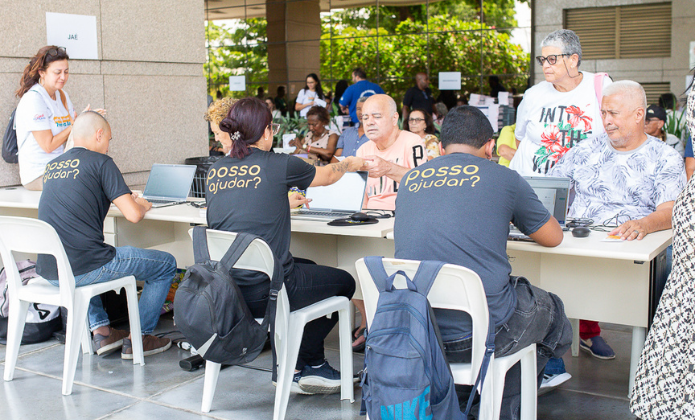 Imagem mostra idosos sentados em frente a uma mesa e sendo atendidos por pessoas, também sentadas, em frente a eles