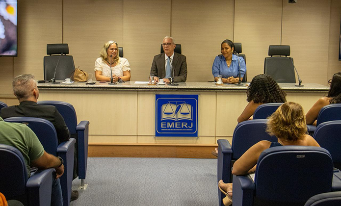 Na foto, vemos uma sala tipo auditório, com paredes de madeira clara ao fundo, com fileiras de cadeiras azuis. Em primeiro plano, aparecem algumas pessoas sentadas de costas para a câmera, compondo a plateia. À frente delas, em mesa comprida e levemente elevada, estão três pessoas sentadas lado a lado, voltadas para o público. No centro, de terno escuro e camisa clara, usando óculos e falando ao microfone está o desembargador Wagner Cinelli. À esquerda dele (para quem olha a foto), está a assistente social Patrícia Valéria Leal trajando blusa de tom claro e sorrindo e com os cabelos claros soltos. À direita, está a colaboradora Alessandra Man-Fu Patrocinio, de cabelo escuro preso, usando uma camisa azul de mangas compridas. Na parte frontal da mesa, há um painel com o logotipo da EMERJ, que inclui uma balança estilizada e a sigla em destaque. As três pessoas na mesa participam de uma palestra dirigida à plateia.