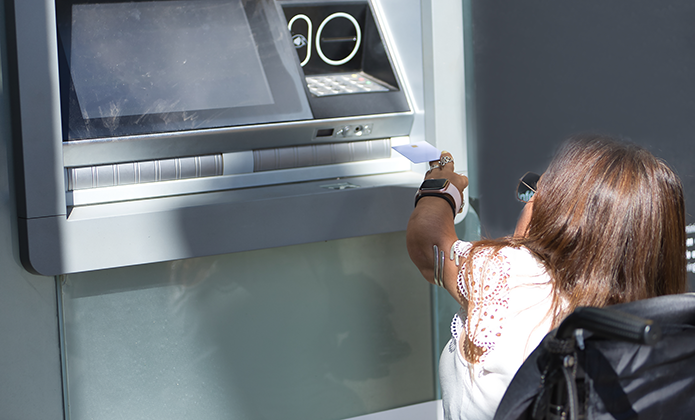 #ParaTodosVerem: A imagem mostra uma mulher utilizando um caixa eletrônico (ATM) ao ar livre. Ela está sentada em uma cadeira de rodas, voltada para a máquina, enquanto insere ou retira um cartão da área de saque. A mulher tem cabelos lisos, castanhos e compridos, usa óculos escuros e uma blusa branca com rendas nas mangas. A máquina de autoatendimento é prateada, moderna e está instalada contra uma parede cinza. O design da estrutura e a posição da mulher sugerem que o caixa não está em altura acessível, pois ela precisa esticar bastante o braço para alcançar a entrada do equipamento. A iluminação é forte, vinda da luz solar direta, indicando que a cena ocorre durante o dia. A imagem chama atenção para questões de acessibilidade e inclusão no uso de serviços bancários por pessoas com deficiência física.