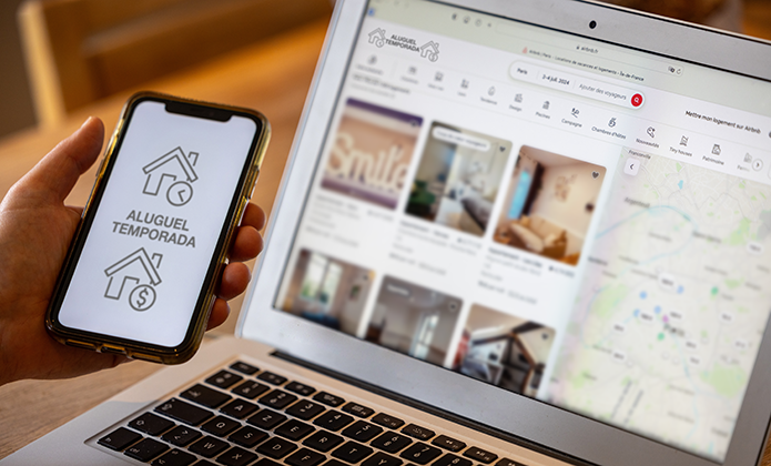 #ParaTodosVerem: Uma pessoa segura um celular que exibe a tela com o texto “Aluguel Temporada” e o ícone de uma casa. Ao fundo, sobre a mesa, está um notebook aberto em um site de buscas de imóveis, mostrando fotos de quartos e um mapa de localização.