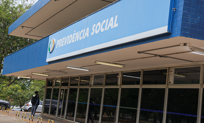 #ParaTodosVerem: Fachada de uma agência da Previdência Social, com estrutura em tons de azul e branco. Na parte superior, uma grande placa exibe o nome “Previdência Social” em letras brancas sobre fundo azul. Abaixo, há uma sequência de portas e janelas de vidro refletindo a área externa. Ao lado da entrada, vê-se uma pessoa caminhando pela calçada. Árvores e vegetação aparecem ao redor, compondo um ambiente urbano e institucional.