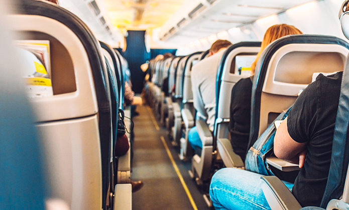 #ParaTodosVerem: Interior de um avião visto pelo corredor central. Passageiros estão sentados em suas poltronas, alguns com roupas casuais, enquanto a luz natural entra pelas janelas. O ambiente é iluminado e mostra a cabine ao fundo.