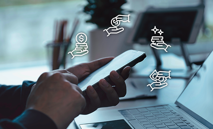 #ParaTodosVerem: A imagem mostra uma pessoa segurando e manuseando um smartphone com as duas mãos, enquanto um laptop está aberto à frente sobre uma mesa, sugerindo um ambiente de trabalho ou estudo. Ao redor do celular, há quatro ícones brancos sobrepostos digitalmente, cada um representando uma mão segurando algo: moedas, notas de dinheiro, um pacote ou um conjunto de itens, e um feixe de estrelas ou brilho, simbolizando possivelmente transações financeiras, pagamentos digitais ou serviços. O fundo está desfocado, indicando que o foco principal é o uso do smartphone e os ícones que representam ações digitais ou econômicas.