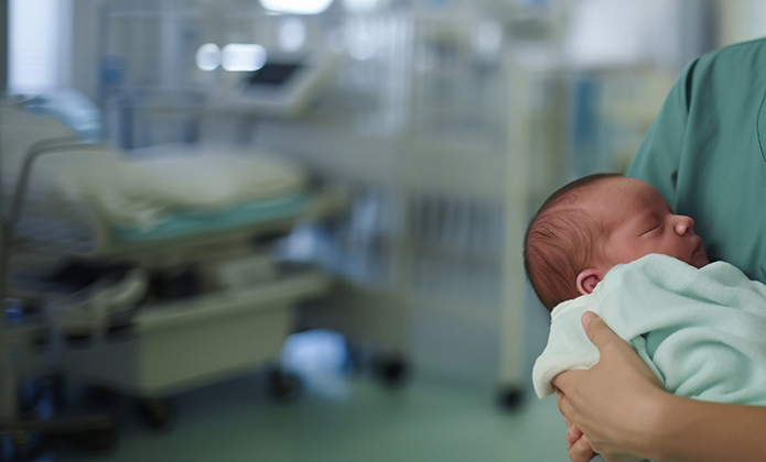 #ParaTodosVerem: Uma profissional de saúde, vestida com roupa hospitalar verde, segura um recém-nascido enrolado em um cobertor claro. Ao fundo, há camas e equipamentos médicos desfocados, indicando que estão em um ambiente hospitalar.