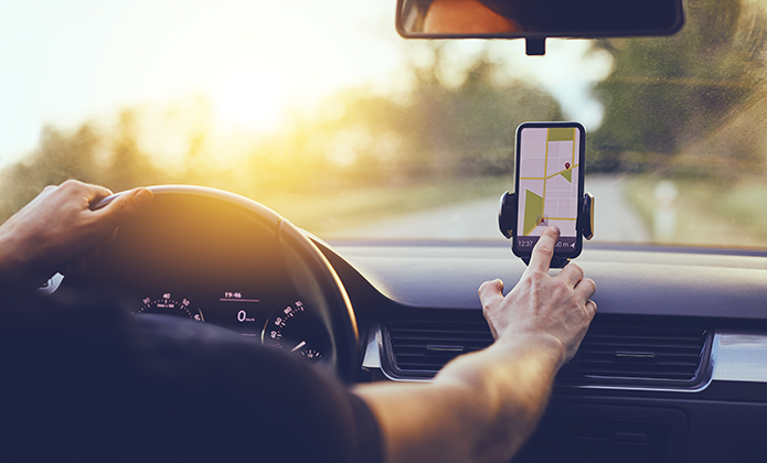 #ParaTodosVerem: Pessoa dirige um carro durante o dia e interage com um celular preso ao painel, que exibe um aplicativo de navegação com mapa e rota.