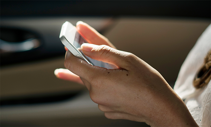 #ParaTodosVerem: Mãos femininas seguram um celular prateado em um ambiente interno, com fundo desfocado que parece ser o interior de um carro. A luz natural ilumina parcialmente as mãos e o aparelho.
