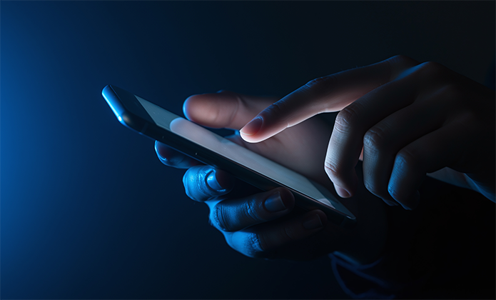 #ParaTodosVerem: Imagem em close mostra mãos usando um celular em ambiente escuro, iluminado pela luz da tela.