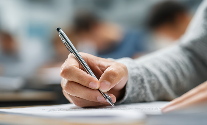 #ParaTodosVerem: Imagem em close mostra mão segurando uma caneta e escrevendo em folha de papel sobre uma mesa; ao fundo, pessoas aparecem desfocadas, sugerindo ambiente de sala de aula ou prova.