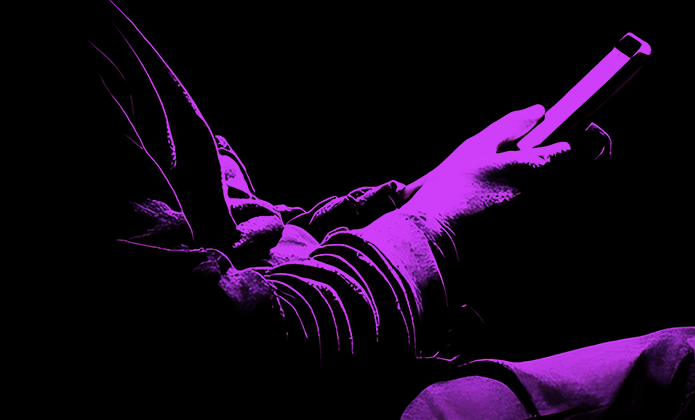 #ParaTodosVerem: Pessoa sentada em ambiente escuro segura um celular nas mãos. A imagem está iluminada com luz roxa intensa, destacando apenas o contorno do corpo e do aparelho contra o fundo preto.