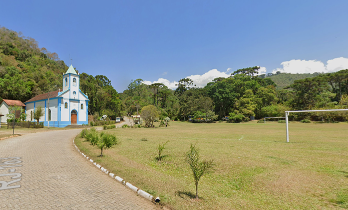 #ParaTodosVerem: Imagem de uma pequena igreja azul e branca à esquerda, com uma torre e cruz no topo, localizada ao lado de uma estrada de paralelepípedos. Ao redor há árvores e vegetação, e à direita um campo gramado com traves de futebol. Ao fundo, morros cobertos por mata e céu azul com poucas nuvens.