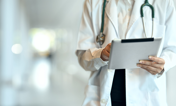 #ParaTodosVerem: Profissional da área da saúde usando jaleco branco e estetoscópio no pescoço segura um tablet, em um ambiente interno iluminado e desfocado ao fundo.