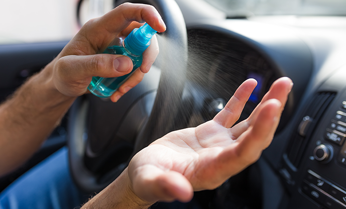 #ParaTodosVerem: A imagem mostra uma pessoa dentro de um carro, borrifando um líquido nas mãos a partir de um frasco spray pequeno e azul. A mão direita está aberta, recebendo o jato, enquanto a esquerda segura o frasco e aciona o gatilho. O volante e parte do painel do veículo estão visíveis ao fundo, sugerindo que a ação ocorre antes ou durante a condução, provavelmente como forma de higienização.