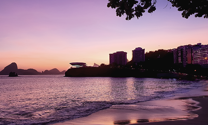 #ParaTodosVerem: Vista panorâmica da Praia de Icaraí, em Niterói, durante o entardecer. O céu exibe tons suaves de rosa, lilás e dourado, refletidos na água calma. À esquerda, as montanhas e o famoso Pão de Açúcar se desenham ao longe. Ao centro, destaca-se o Museu de Arte Contemporânea (MAC), com sua forma circular futurista. À direita, a orla urbana é marcada por edifícios altos, iluminados por luzes amareladas, e a silhueta de árvores completa a cena. O mar tranquilo avança levemente sobre a faixa de areia em primeiro plano.