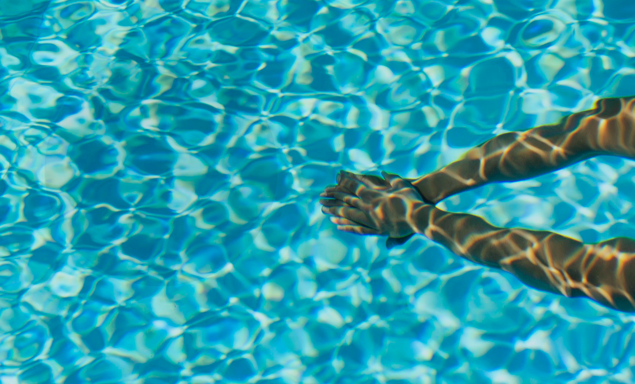 #ParaTodosVerem: A imagem mostra uma piscina com água cristalina em tons de azul, refletindo a luz do sol que cria padrões ondulados e brilhantes na superfície e no fundo da água. No lado direito da imagem, há dois braços estendidos, com as mãos unidas em posição de mergulho, sugerindo que a pessoa está prestes a mergulhar ou nadar. A luz do sol projeta sombras e reflexos nos braços, criando um efeito visual interessante de faixas de luz.