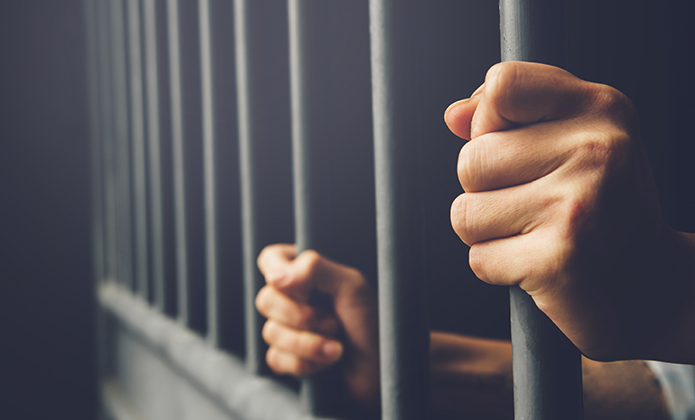 #ParaTodosVerem: Imagem em close mostra duas mãos de homem branco segurando firmemente as grades de uma cela, com fundo escuro e iluminação lateral.