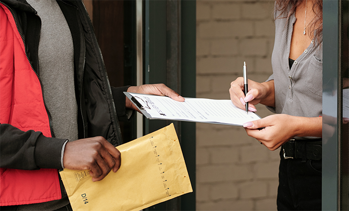 #ParaTodosVerem: A imagem mostra uma mulher e um entregador durante o processo de recebimento de uma encomenda. O entregador, vestindo colete vermelho e casaco escuro, segura um envelope grande de papel pardo enquanto a mulher, parcialmente visível, assina um formulário de recebimento sobre uma prancheta. A cena ocorre diante de uma porta, sugerindo a entrada de uma residência ou escritório.