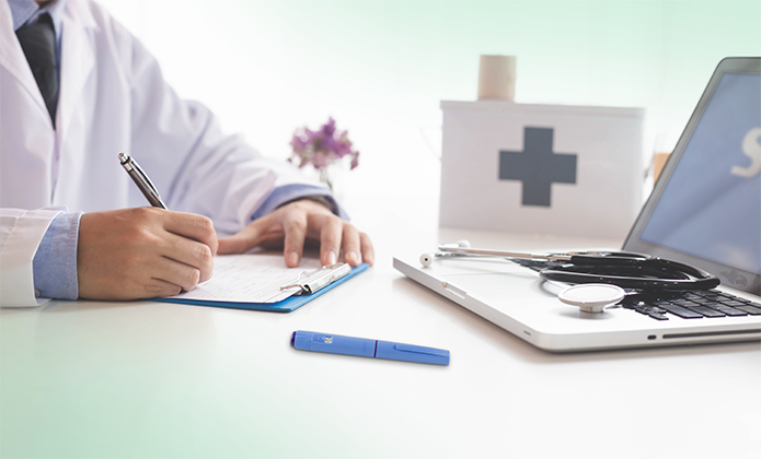 #ParaTodosVerem: A imagem mostra a cena de um consultório médico. Em primeiro plano, à esquerda, está a mão de uma pessoa vestindo jaleco branco e camisa azul-clara, segurando uma caneta e escrevendo sobre uma prancheta azul com folhas brancas. Sobre a mesa, em tons claros, há um objeto retangular azul (parece uma caneta-teste ou marcador), um notebook aberto com a tela parcialmente visível e um estetoscópio repousando sobre o teclado. Ao fundo, um recipiente branco com uma cruz cinza-escura estampada (indicando uso médico ou de primeiros socorros) está sobre a mesa, levemente desfocado, e, próximo a ele, há um pequeno arranjo floral roxo. O ambiente é iluminado por luz suave, dando um ar limpo e organizado, típico de um cenário clínico.