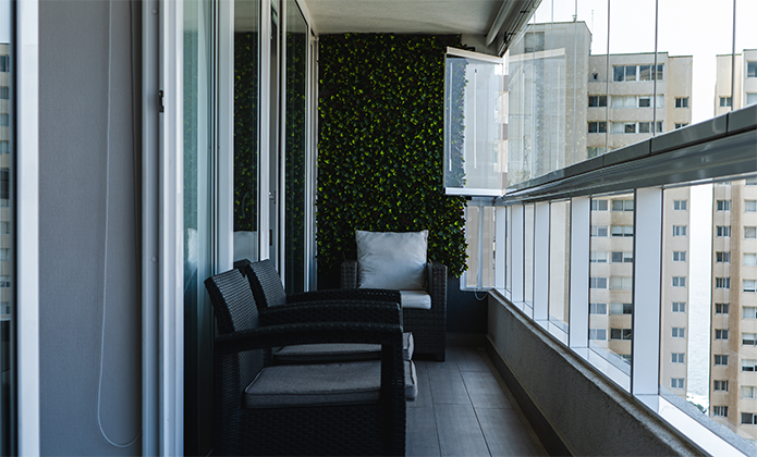 #ParaTodosVerem: Varanda moderna de um apartamento, envidraçada do lado direito, com vista para edifícios altos. À esquerda, há portas de vidro que dão acesso ao interior do apartamento. A varanda é decorada com um painel verde vertical de plantas artificiais ao fundo, diante do qual estão duas poltronas de fibra sintética com almofadas cinza-claro. Uma terceira poltrona e uma pequena mesa de apoio aparecem parcialmente ao fundo. A iluminação é natural, entrando pelas janelas abertas, e o espaço transmite sensação de conforto e organização.