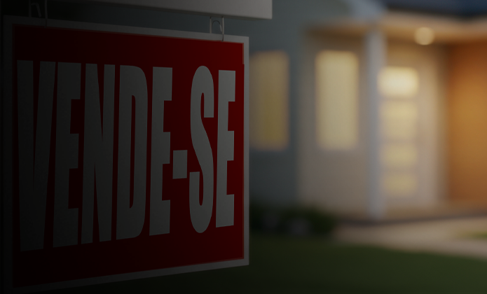 #ParaTodosVerem: A imagem mostra uma placa vermelha com letras brancas em destaque escrito “VENDE-SE”. Ao fundo, aparece uma casa desfocada, sugerindo que o imóvel está à venda.